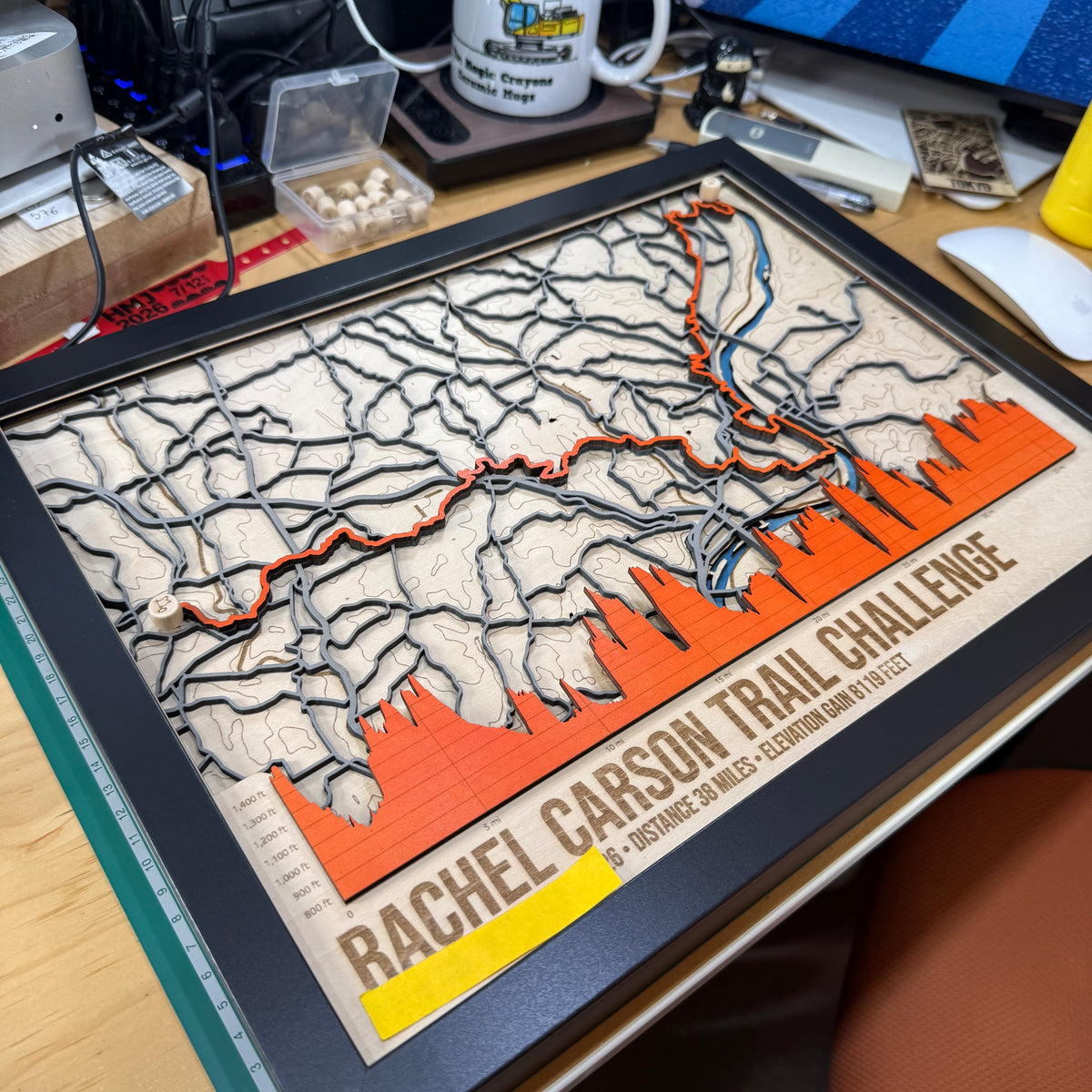 A framed 3D wooden trail map plaque celebrating the Rachel Carson Trail Challenge, featuring a topographic map design with an orange profile chart and text at the bottom, displayed on a desk.